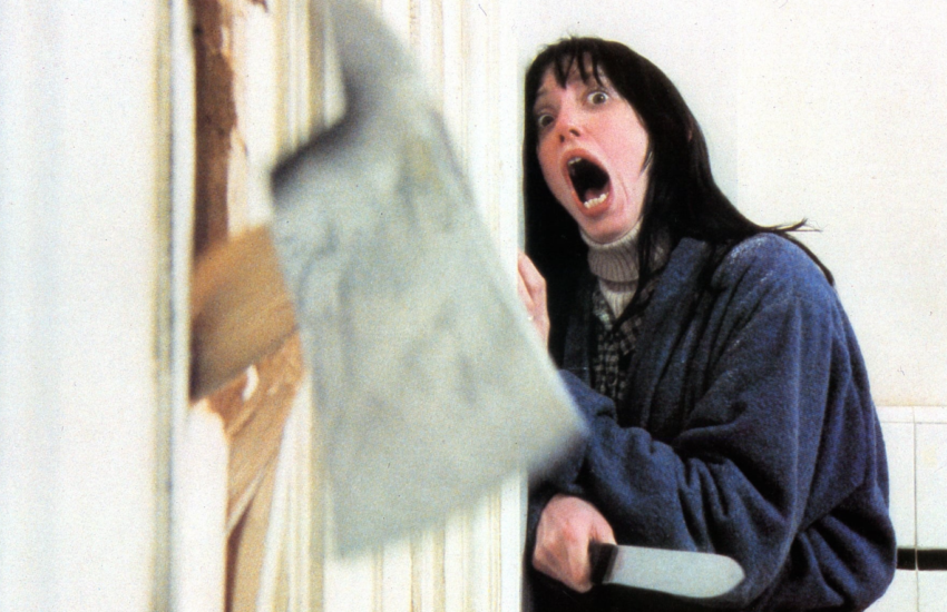 Woman leaning against the wall, screaming at an ax that has cut through from the next room